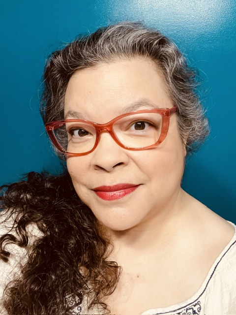Lisa, a Latina with dark hair going gray, stands against a teal colored wall. She wears red glasses and lipstick.